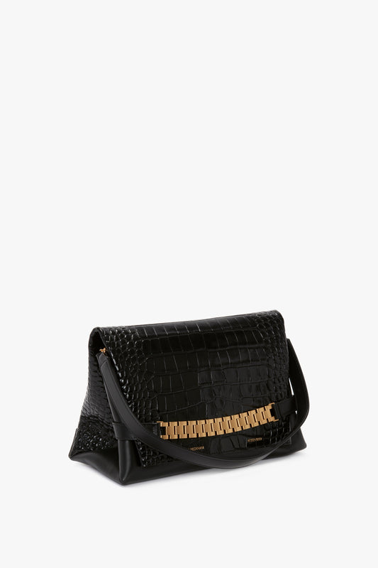 A black leather handbag with a crocodile skin pattern and a gold chain detail, featuring a shoulder strap, is displayed against a white background. This elegant piece by Victoria Beckham showcases luxurious croc embossed leather, making it an ideal Chain Pouch Bag With Strap In Black Croc-Effect Leather for sophisticated outings.