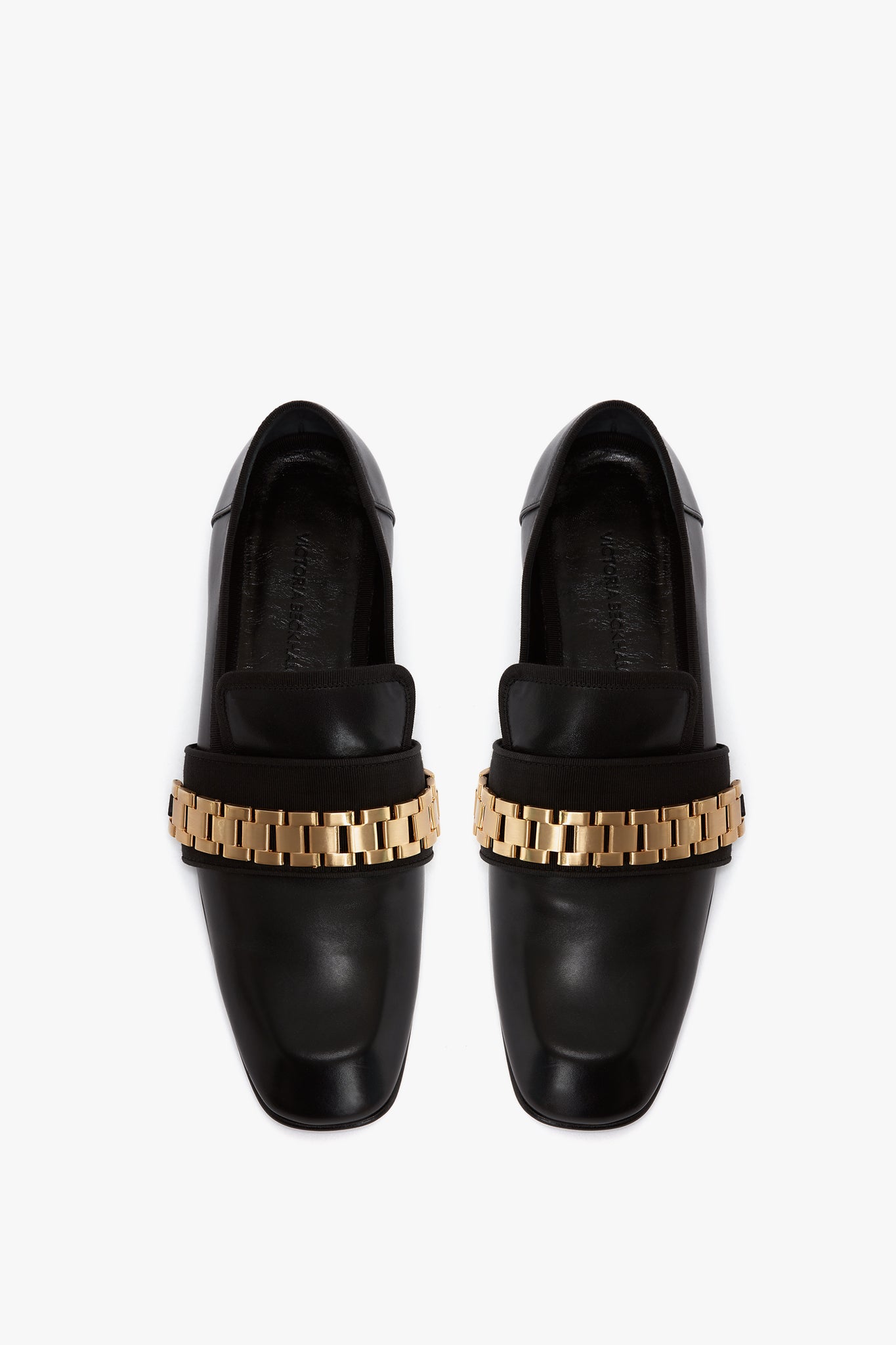 A pair of black patent leather Mila Chain Loafer In Black by Victoria Beckham with gold chain detailing across the top, viewed from above against a white background.