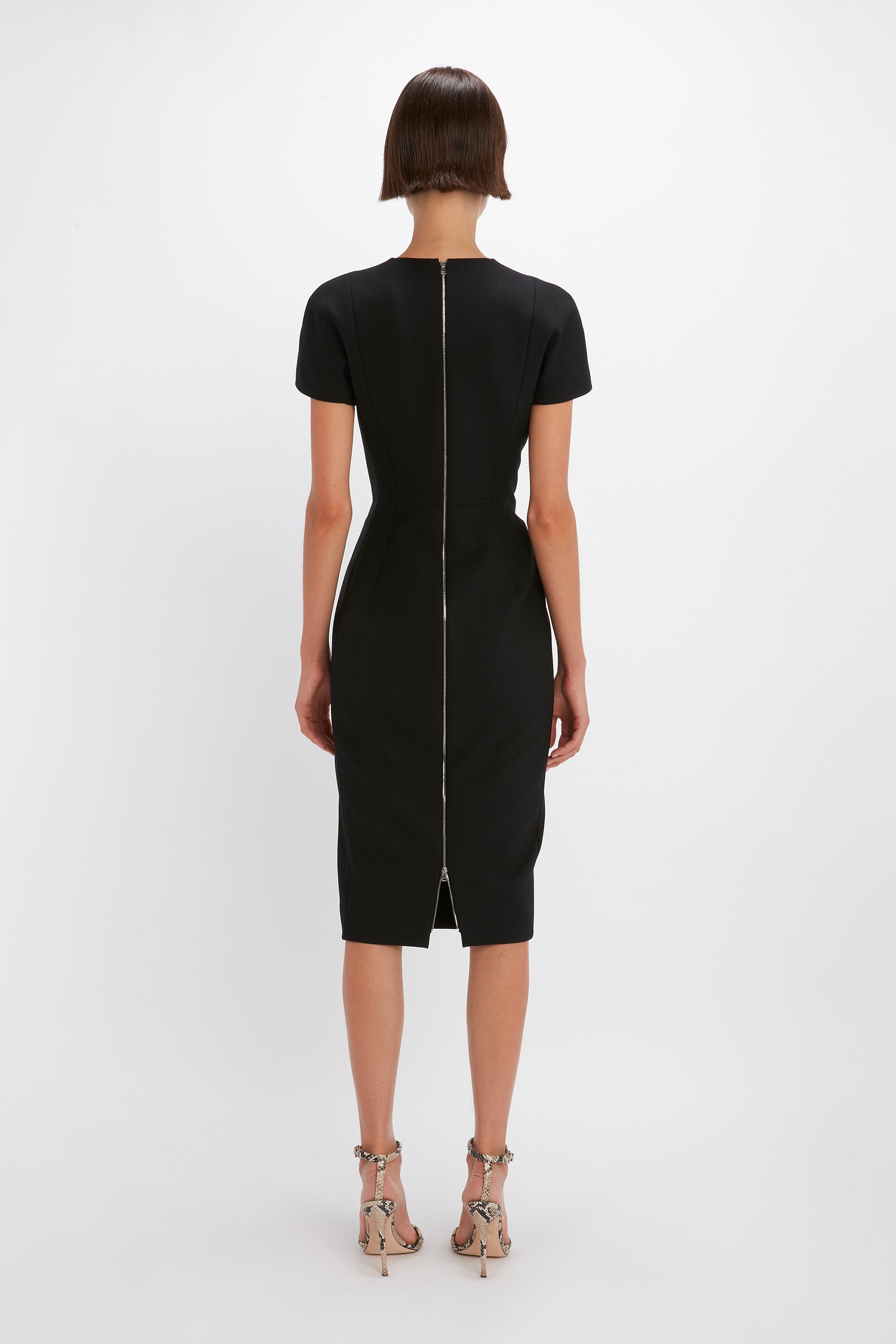 A person with short hair is standing with their back facing the camera, wearing a knee-length, short-sleeved black Spiral Fitted T-Shirt Dress In Black by Victoria Beckham with a visible back zipper. The elegant silhouette is complemented by high-heeled sandals.