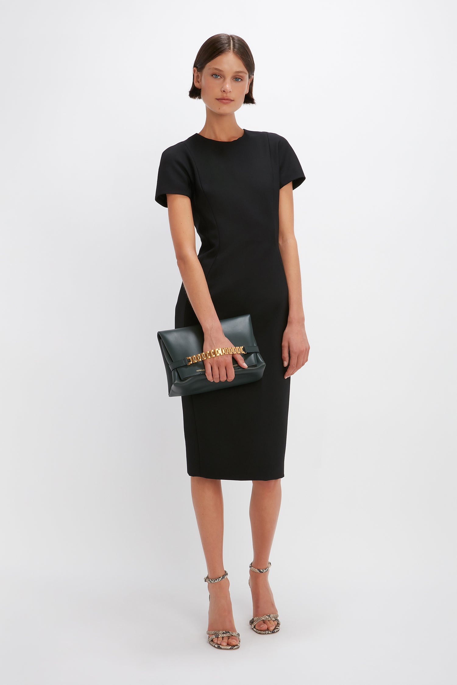 Person standing against a plain background, wearing the Victoria Beckham Spiral Fitted T-Shirt Dress In Black and holding a black clutch with a gold chain. They have short hair and are wearing strappy heels, embodying a sleek Victoria Beckham style.