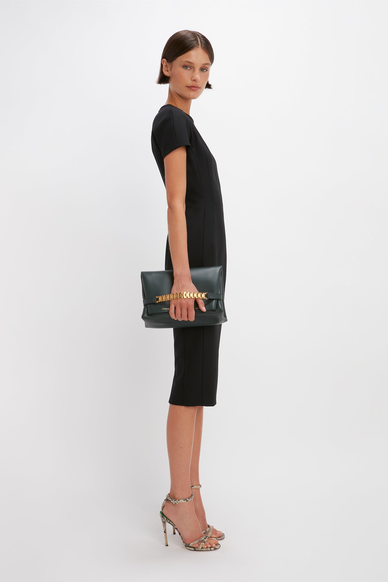 A person in a Victoria Beckham Spiral Fitted T-Shirt Dress In Black, holding a black clutch bag and wearing high-heeled sandals, stands against a plain white background.