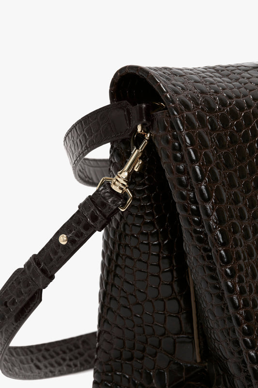 Close-up of a black, textured handbag with a croc-effect Chocolate Brown leather pattern, featuring a metal clasp connecting the strap to the bag. This accessory ensures versatile styling for any occasion. The Jumbo Chain Pouch Bag In Chocolate Croc-Effect Leather by Victoria Beckham is perfect for any occasion.
