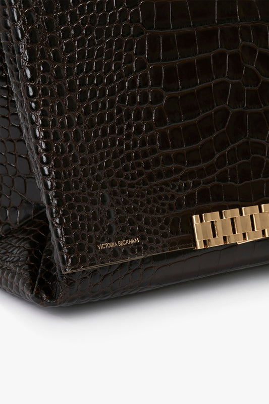 Close-up of a textured, croc-effect Chocolate Brown Jumbo Chain Pouch Bag In Chocolate Croc-Effect Leather with a gold clasp and "Victoria Beckham" branding in small gold lettering, perfect for versatile styling.
