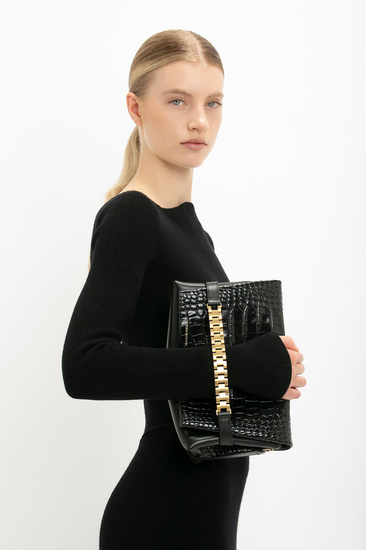 Person in a black outfit holding a black and gold Victoria Beckham Chain Pouch Bag With Strap In Black Croc-Effect Leather against a white background.