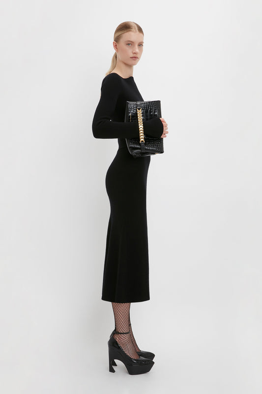A woman stands in profile, exuding Victoria Beckham's signature elegance, wearing a long black dress, fishnet stockings, and high-heeled shoes. She holds a chic Chain Pouch Bag With Strap In Black Croc-Effect Leather by Victoria Beckham. The background is plain white.