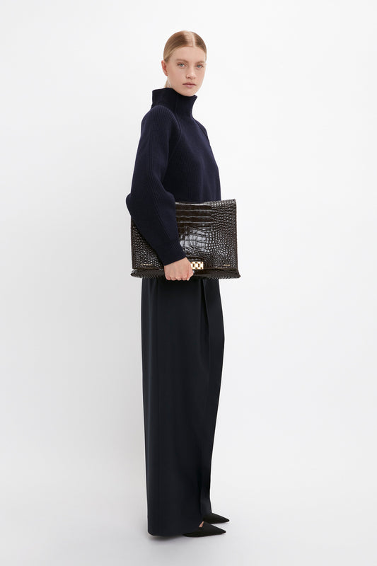 A person stands against a white background, wearing dark wide-leg trousers and a navy turtleneck sweater, holding a Victoria Beckham Jumbo Chain Pouch Bag In Chocolate Croc-Effect Leather, showcasing versatile styling.