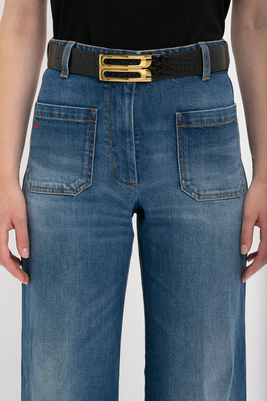 Person wearing high-waisted blue jeans with front pockets and a black Jumbo Frame Belt In Chocolate Croc-Effect Leather by Victoria Beckham.